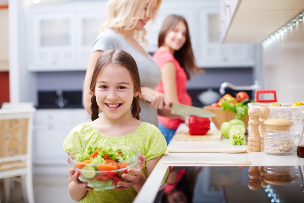 Criança com pote de salada ao fundo desfocado mãe e irmã cortando saladas